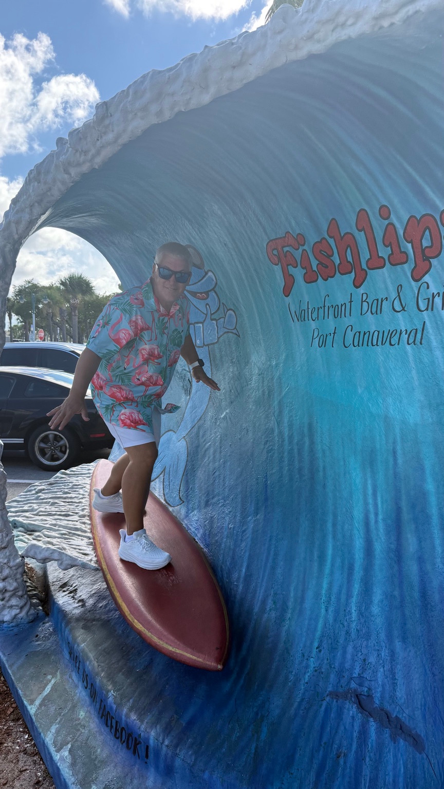 Brad wearing sunglasses and a flamingo shirt, posing on a surfboard display inside a large sculpted blue wave at Fishlips